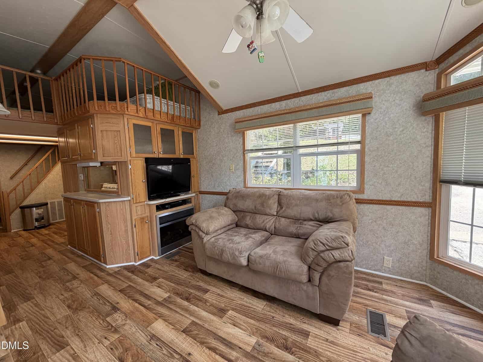 A cozy living room with a brown couch, wood floors, large window, TV stand, and loft area with wooden railing. Stairs and kitchen are visible in the background. Ceiling fan hangs overhead.