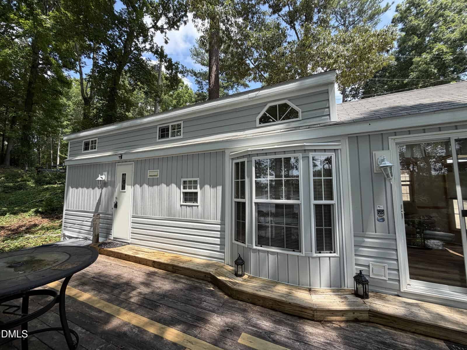A light gray house with white trim, large windows, and a wooden deck surrounded by trees. A round metal table sits on the deck, and small lanterns are near the door and windows.
