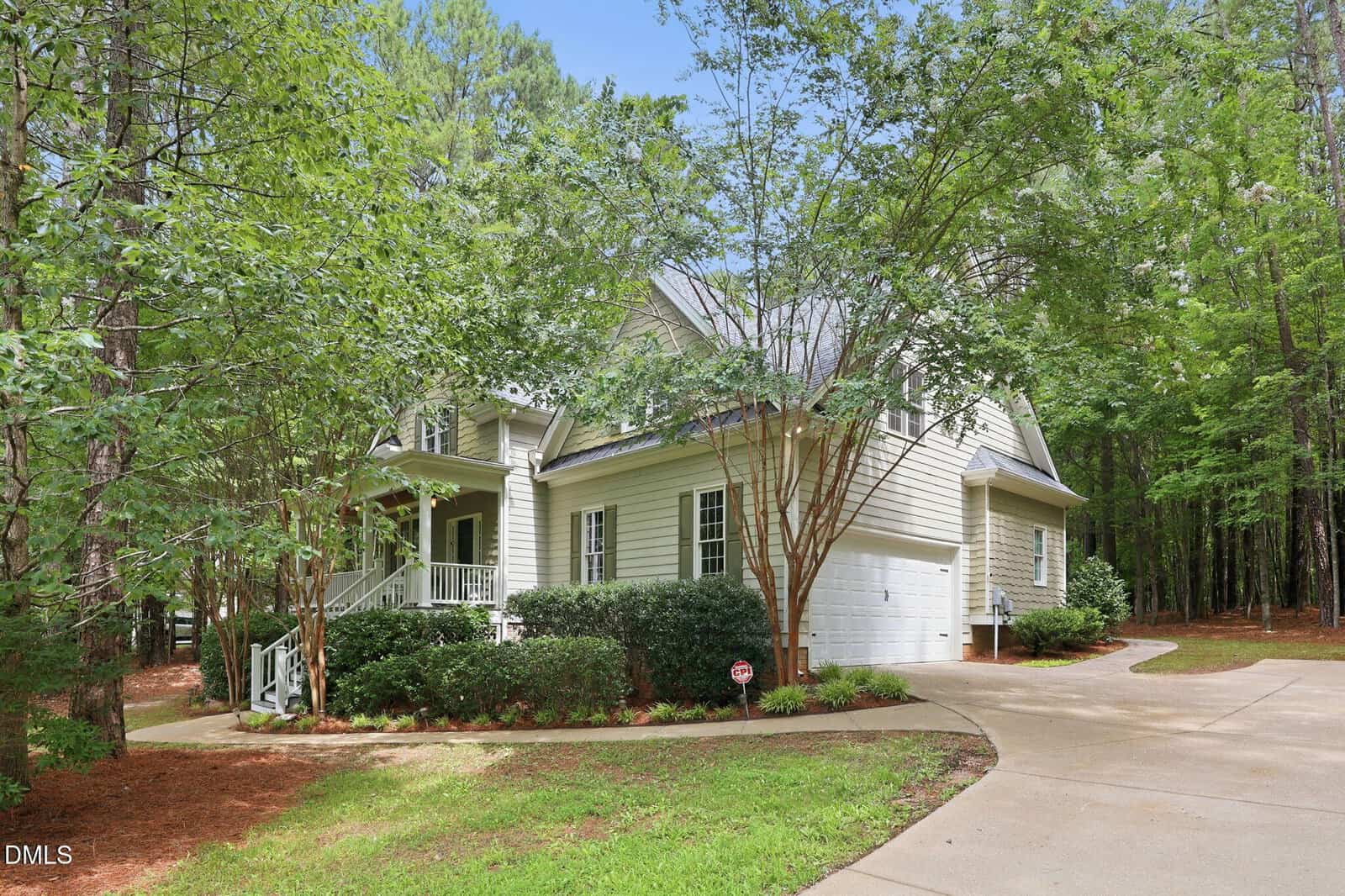 Two-story white house with a front porch, surrounded by tall trees and shrubs. A curved driveway leads to a two-car garage. The yard is landscaped and bordered by forest.