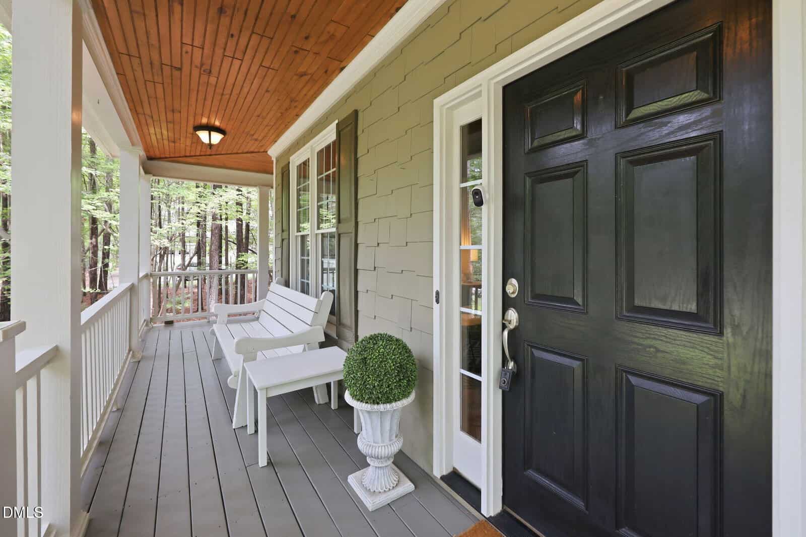 A covered porch with a black front door, white bench seating, a potted plant, wooden ceiling, and railing overlooks trees and greenery.