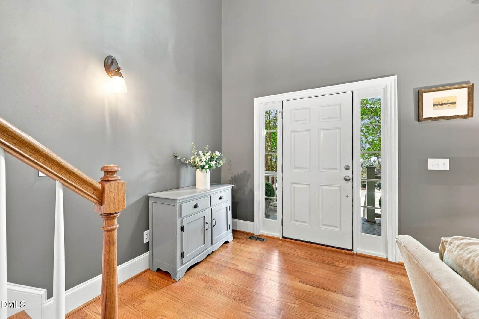 Bright foyer with hardwood floors, white front door with sidelights, gray console table topped with a flower vase, wall sconce above, and staircase with wooden handrail on the left.