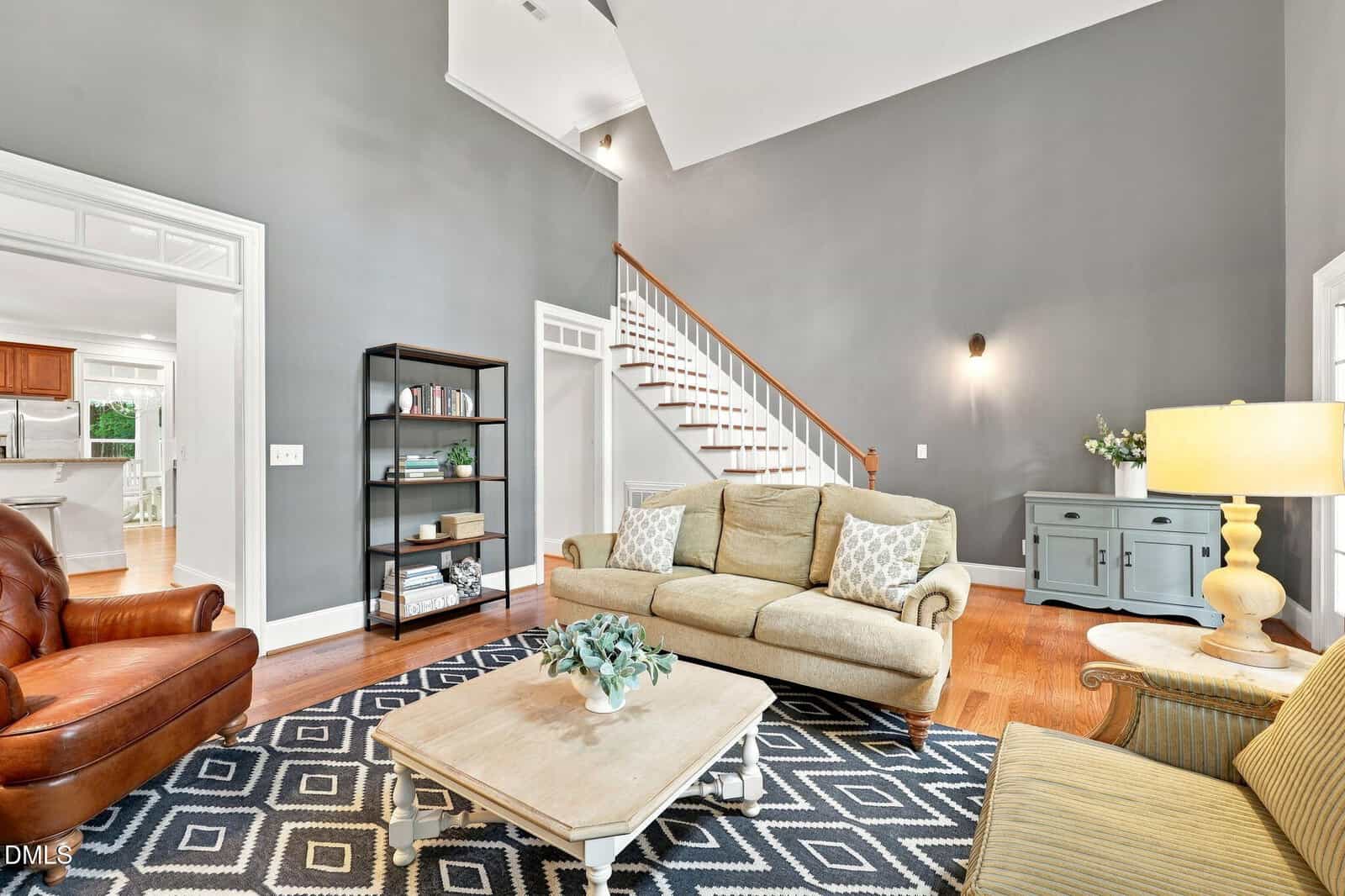 A bright living room with gray walls, a beige sofa, leather chair, patterned rug, coffee table, bookshelf, sideboard, staircase, and wood floors, with a glimpse of a white kitchen in the background.
