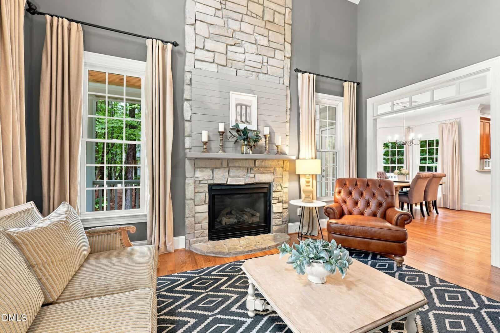 A bright living room with a stone fireplace, tan sofa, brown leather armchair, geometric rug, and large windows with curtains. The room opens to a dining area with a wooden table and chairs.