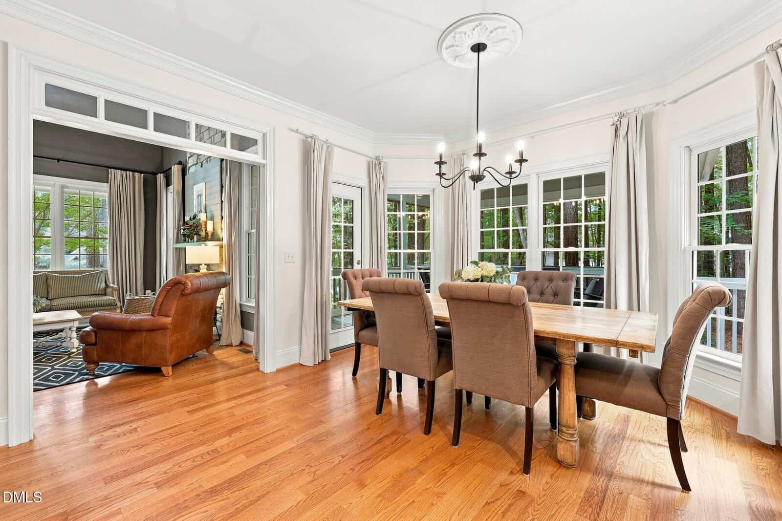 Bright dining room with a wooden table, six upholstered chairs, a black chandelier, large windows with beige curtains, light walls, and hardwood floors, opening to a cozy sitting area with a leather chair.