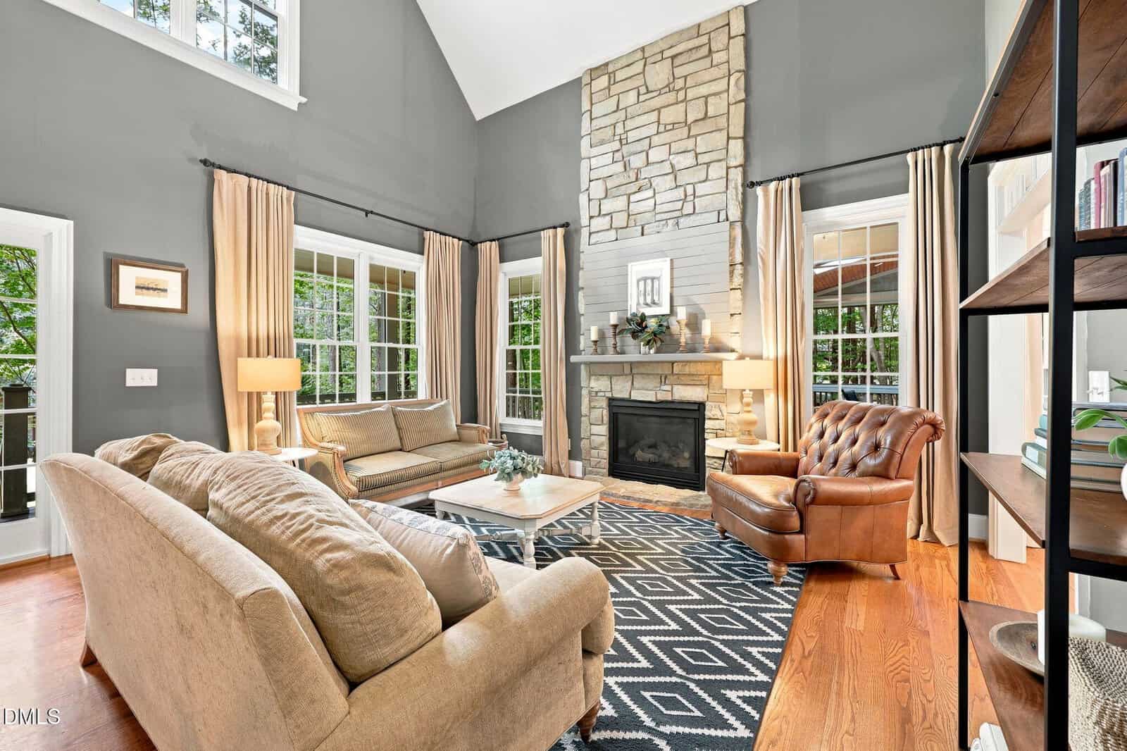 Bright living room with large windows, beige sofas, a brown leather chair, wood floors, a geometric rug, and a stone fireplace. Light curtains frame the windows, and greenery is visible outside.