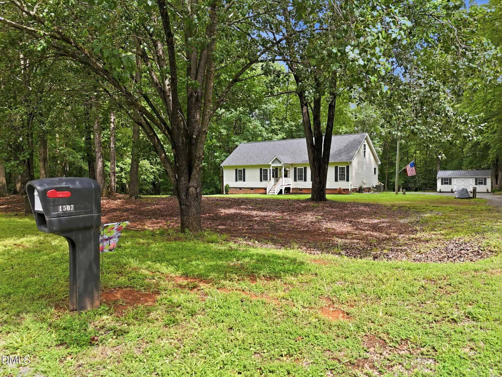 A white house with a front porch sits among large trees at 1507 Rainbow Circle, Oxford, with a black mailbox labeled "1907" in the foreground and a U.S. flag near a detached garage in the background.