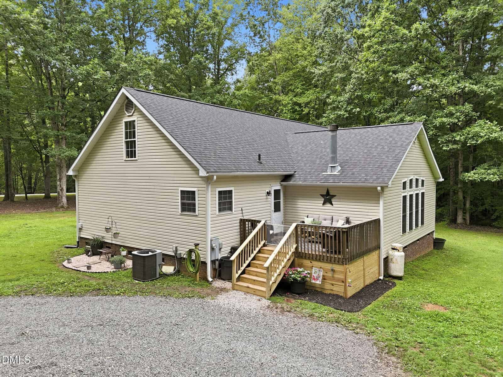 Single-story tan house with gray roof, small wooden deck, wood stove chimney, gravel driveway, and surrounded by green grass and trees under a blue sky.