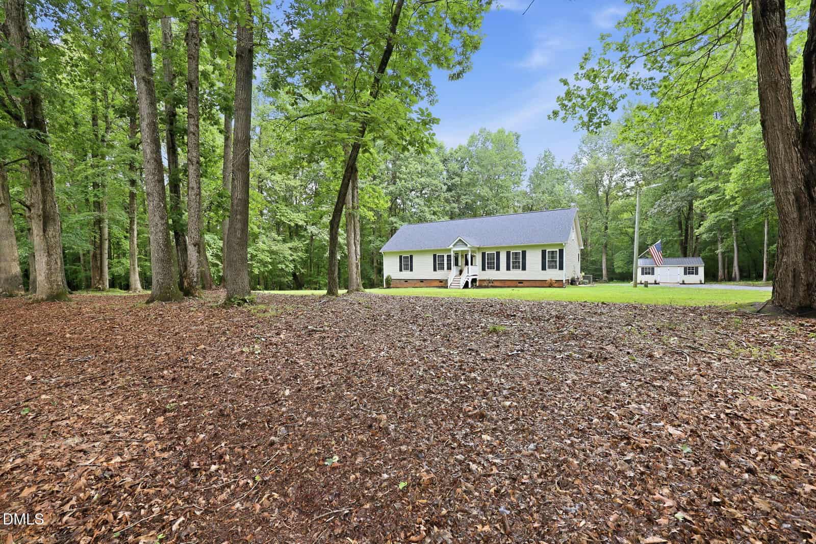 A yellow house with a front porch is set among tall trees, with a large, leaf-covered yard in the foreground. Another small white building is visible in the background to the right.