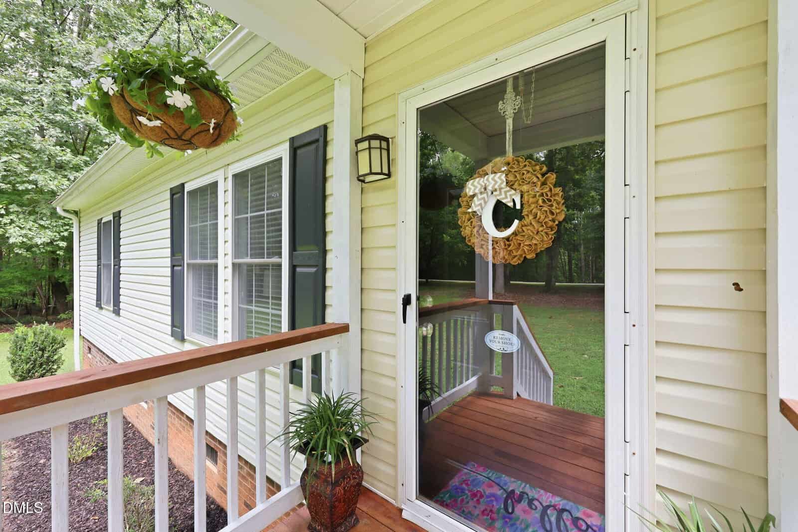 A cozy front porch with a potted plant, a hanging basket of flowers, and a glass door decorated with a wreath. The porch has wooden railings and overlooks a green yard with trees in the background.