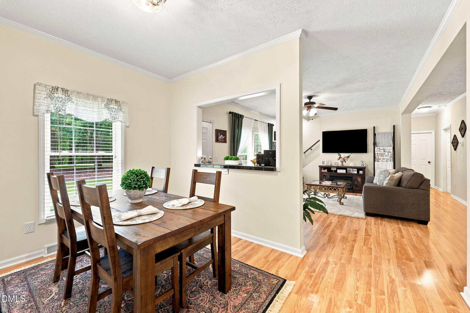 A dining area with a wooden table and four chairs is next to a window; it opens to a living room with a sofa, TV, and ceiling fan. Hardwood floors and light walls create a bright, welcoming atmosphere.