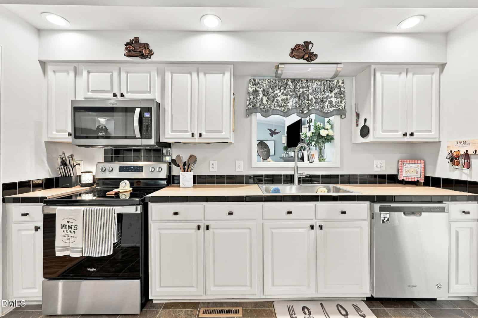 A modern kitchen with white cabinets, black countertops, stainless steel appliances, a window with a floral valance, and various utensils and decor neatly arranged.