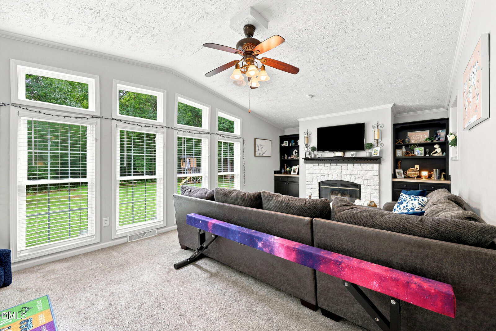 A bright living room with large windows, gray sectional sofa, ceiling fan, white brick fireplace with TV above, built-in shelves, and a colorful purple-pink gymnastics beam behind the couch.