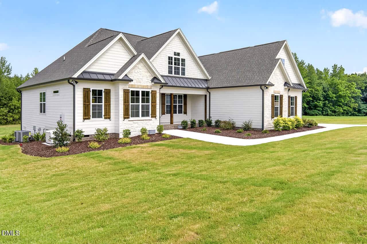 A modern white single-story house with dark roof, large windows, and wooden shutters sits on a spacious green lawn with landscaped shrubs and a concrete driveway under a blue sky.