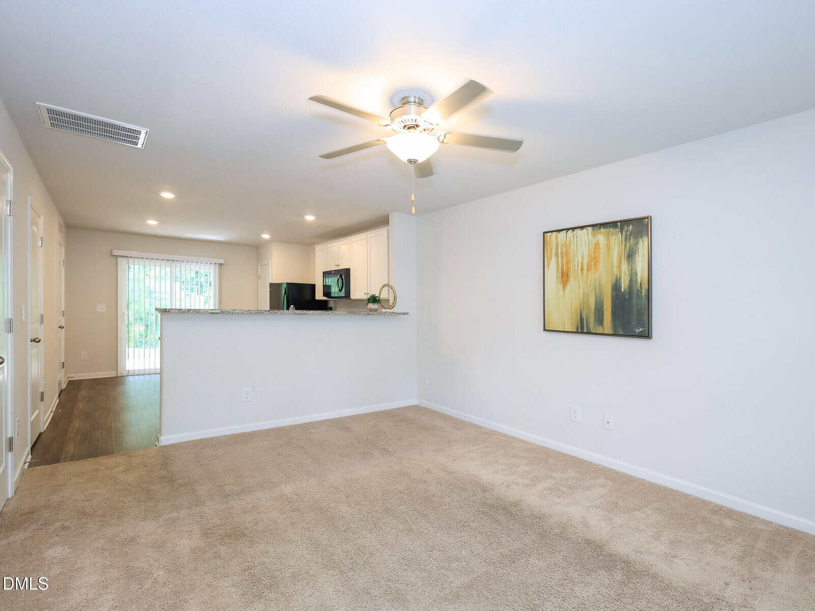 A spacious living room with beige carpet, ceiling fan, abstract art on the wall, and an open view of the kitchen with white cabinets. A sliding door leads to the outside. Neutral colors throughout.