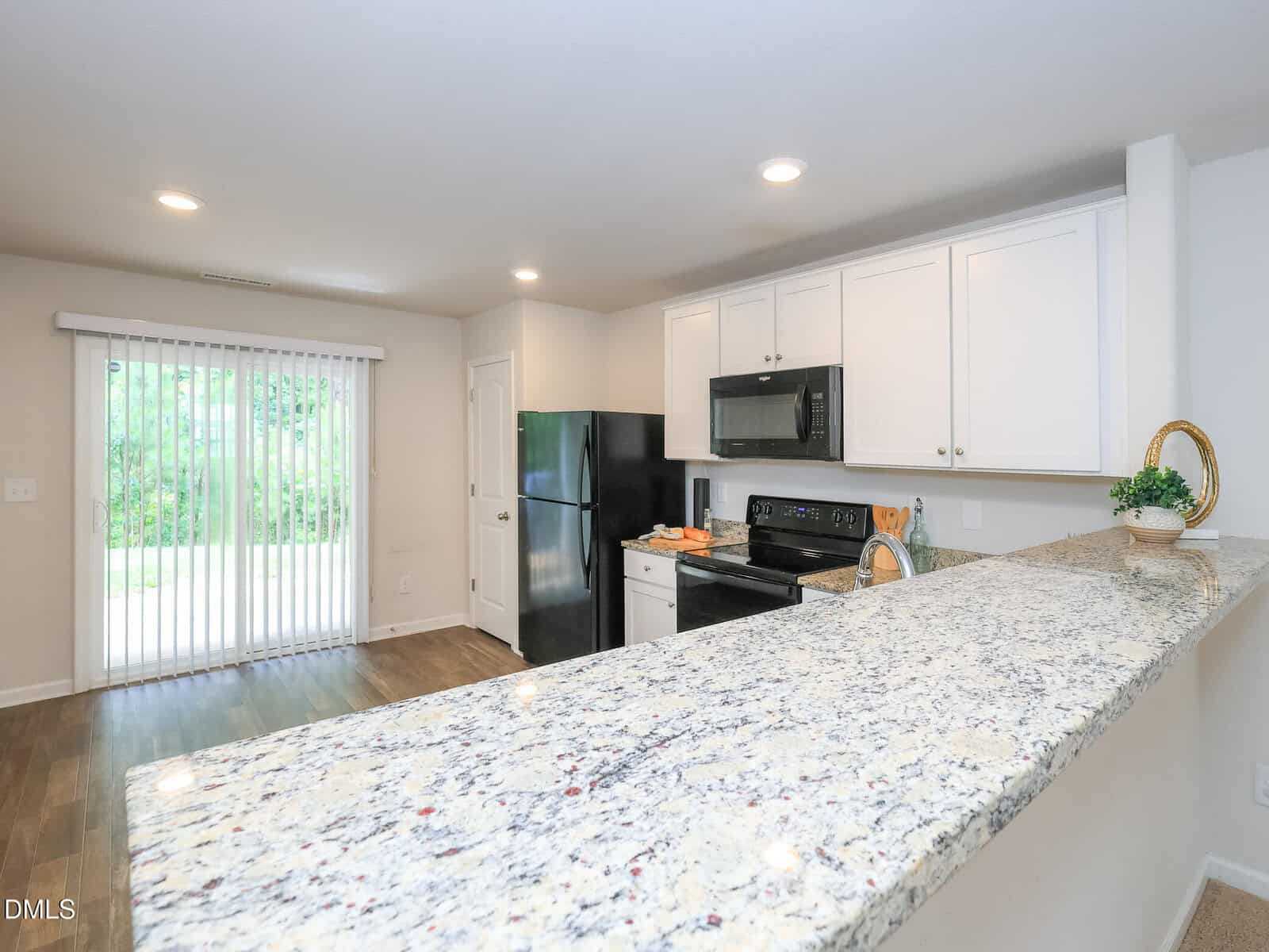 Modern kitchen with granite countertop, white cabinets, black appliances, and wooden floors. Sliding glass doors lead to a green outdoor area, allowing natural light into the space.