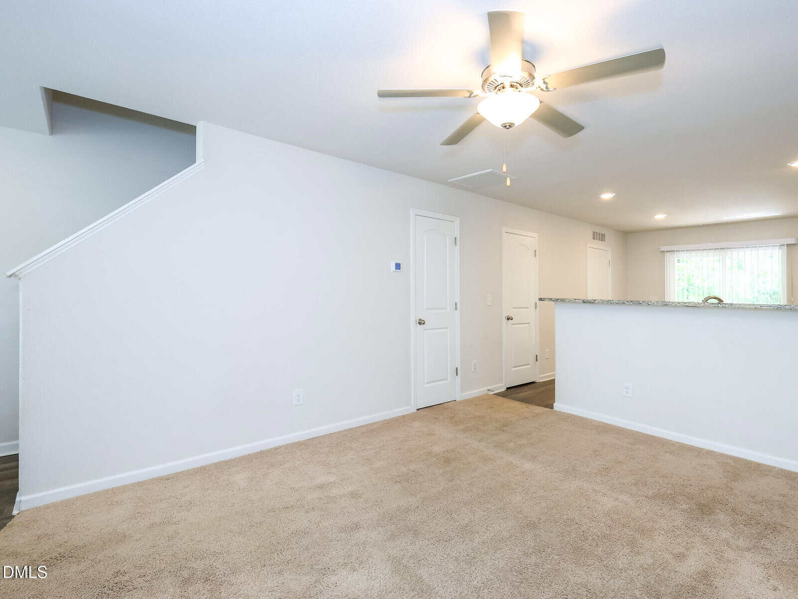 Bright living room with beige carpet, white walls, ceiling fan with light, and partial view of a stairway, hallway door, and granite kitchen counter. Sliding glass doors let in natural light at the back.