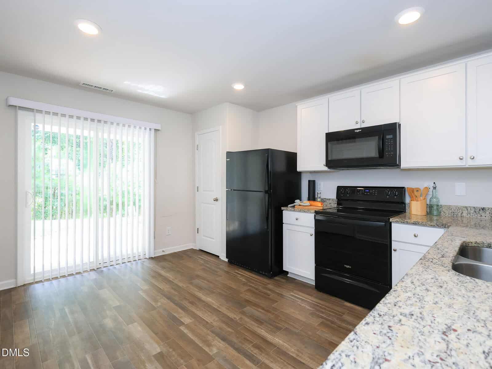 Modern kitchen with white cabinets, black appliances, granite countertops, and wood flooring. Sliding glass doors on the left let in natural light and offer a view of a green outdoor area.