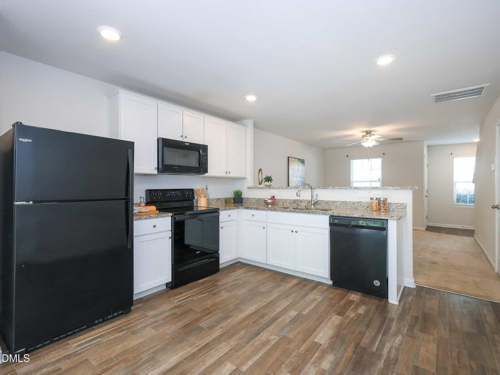 Modern kitchen with white cabinets, black appliances, granite countertops, and wood flooring. An open doorway leads to a bright living area with a ceiling fan and windows in the background.
