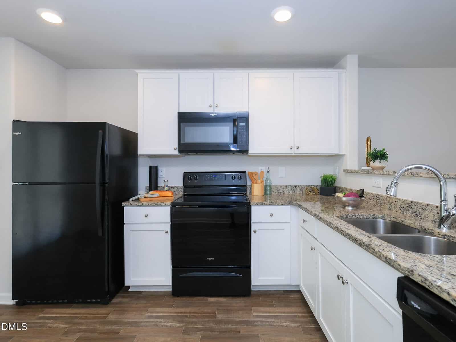 Modern kitchen with white cabinets, black appliances, granite countertops, a double sink, wooden floor, and recessed lighting. Cooking utensils and decorative items are on the countertops.