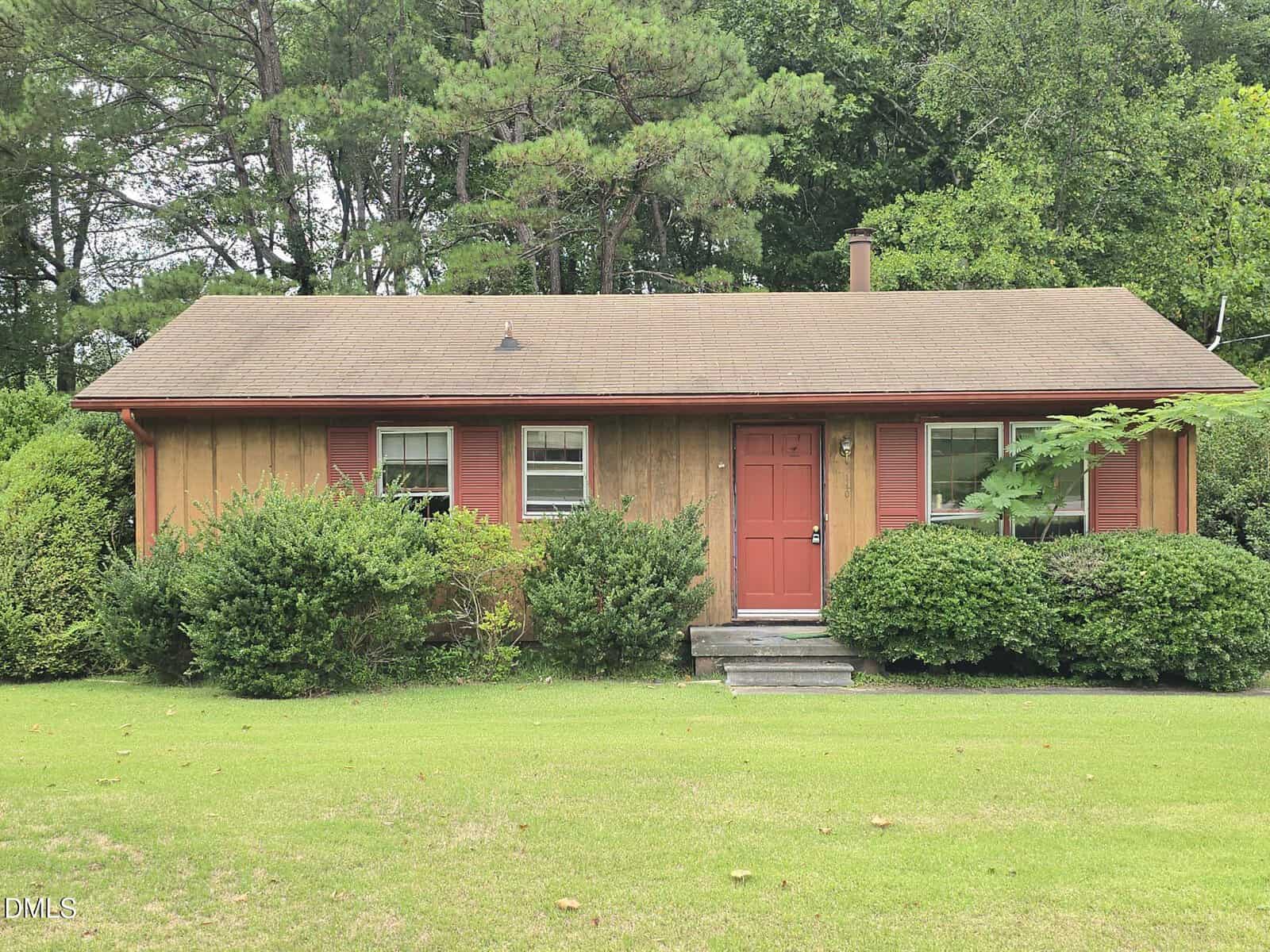 A small, single-story wooden house with a red door, red shutters, and a brown roof sits at 140 Evans Road in Henderson, surrounded by green bushes and trees, with a well-kept lawn in front.