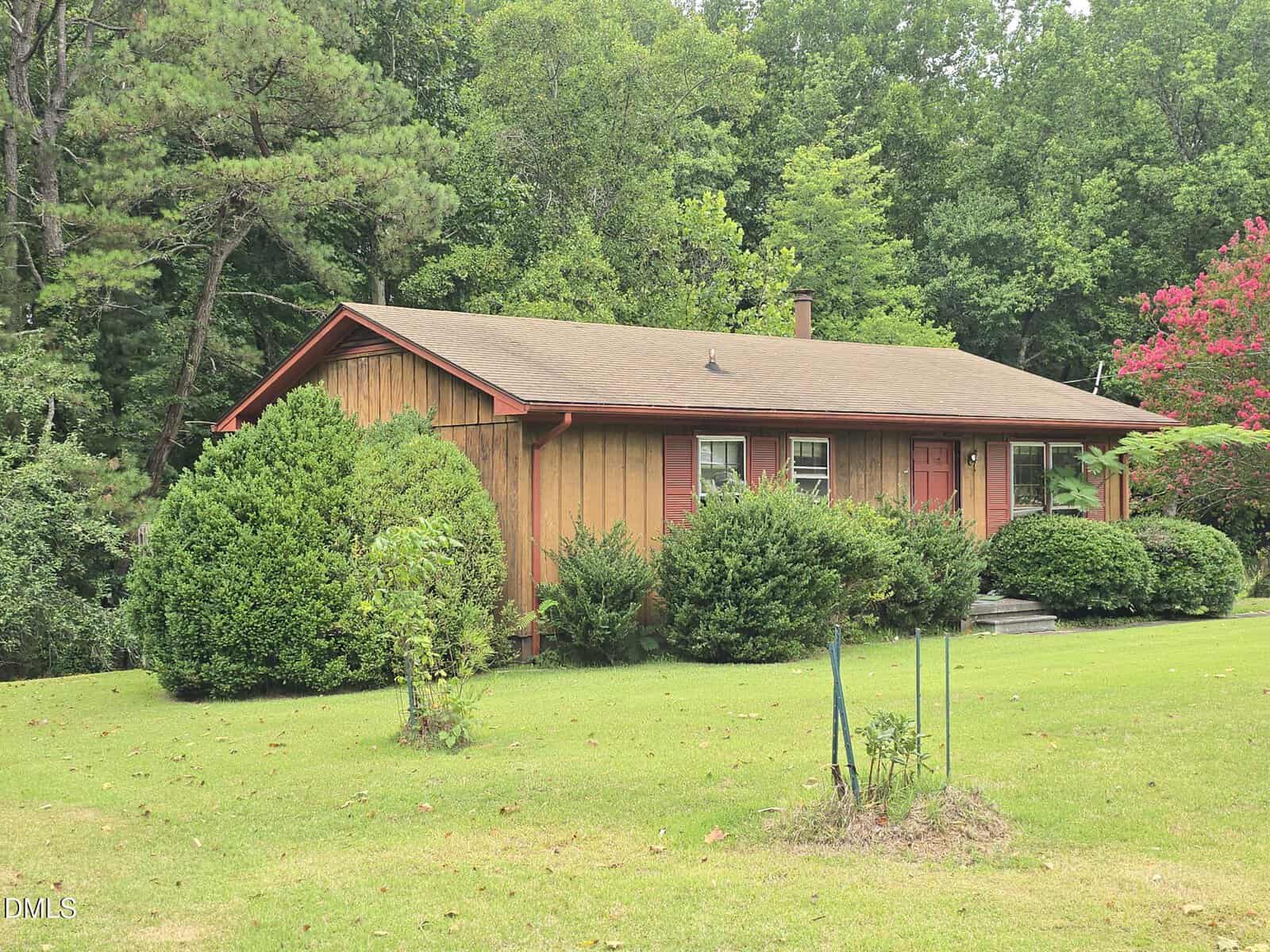 A small, single-story wooden house with a brown roof sits among lush green bushes and trees, with a well-kept lawn in the foreground.