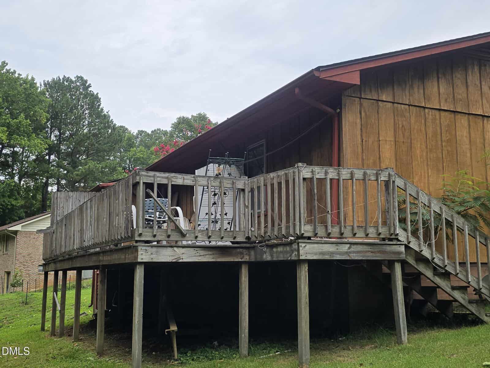 Wooden deck with weathered railings and stairs attached to a brown house. Patio furniture and miscellaneous items are on the deck. The yard below is grassy, with trees and another house in the background.