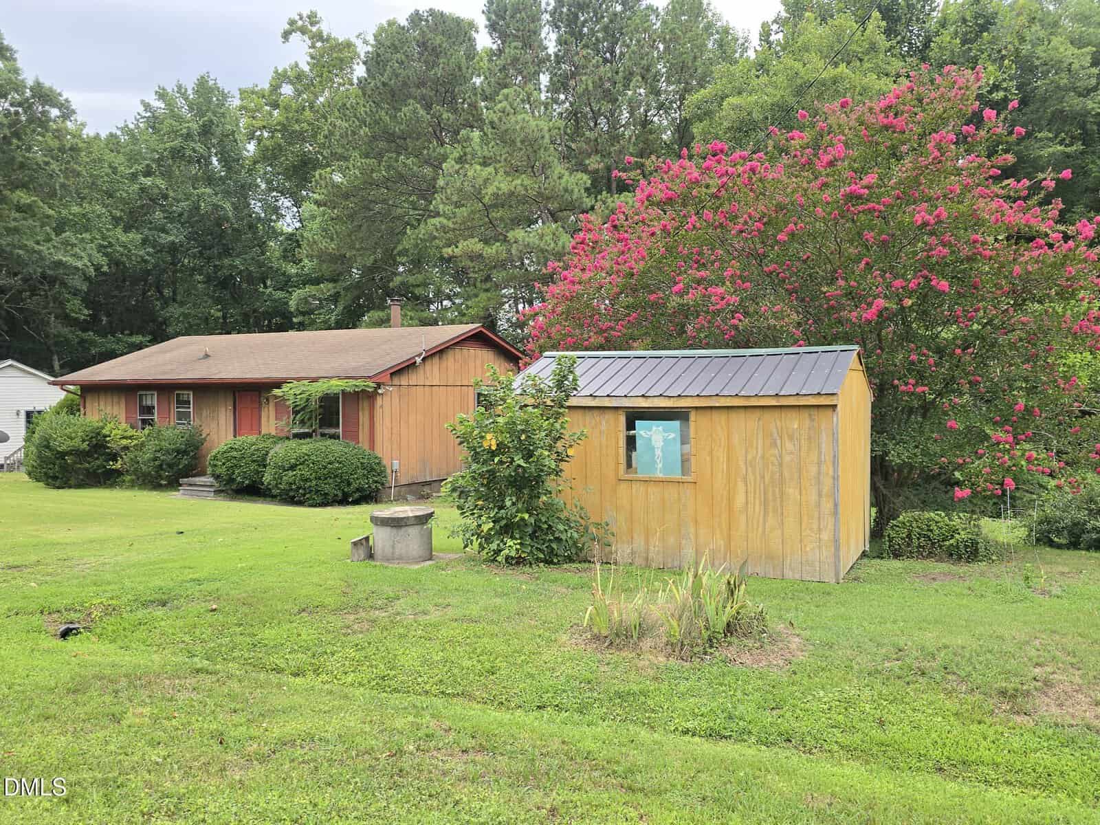 A small wooden house and shed sit on a grassy lawn, surrounded by trees and blooming pink flowers. The sky is overcast.