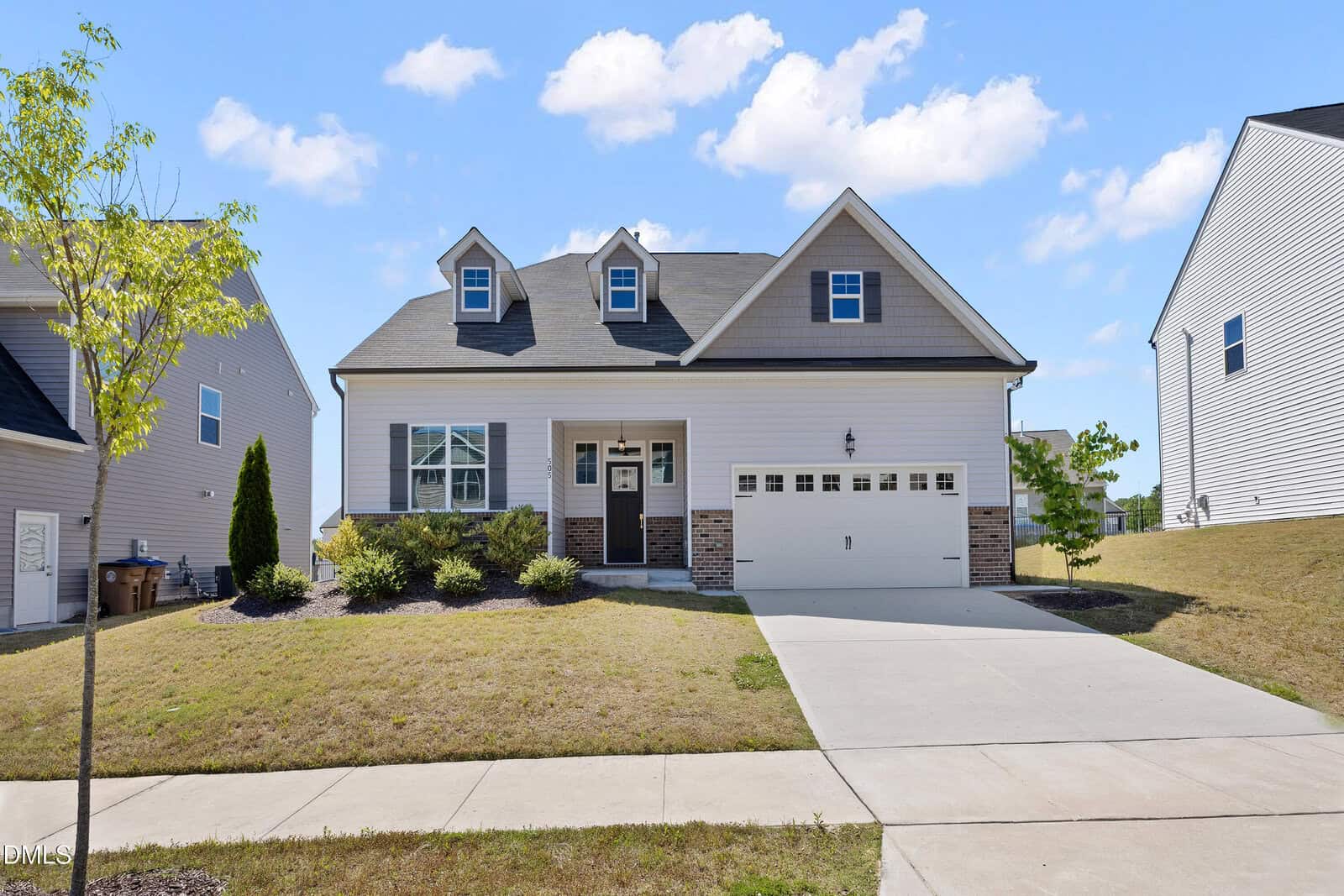 A two-story suburban house at 505 Rain Drizzle Court in Wake Forest, featuring a welcoming front porch, two charming dormer windows, an attached two-car garage, and a sloped lawn with minimal landscaping on a sunny day.