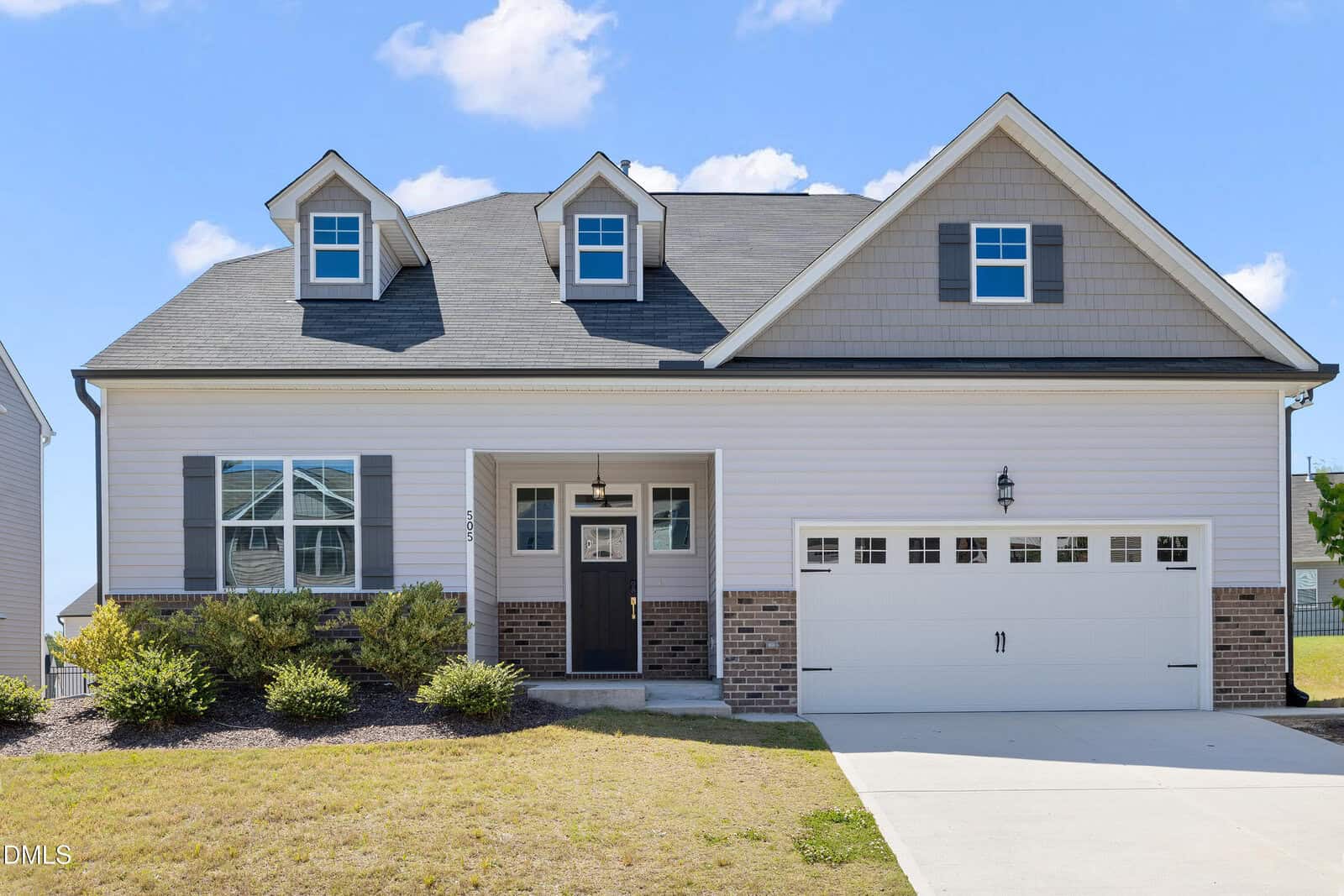 A modern suburban house with gray siding, brick accents, a two-car garage, dormer windows, and a manicured lawn under a blue sky with scattered clouds.