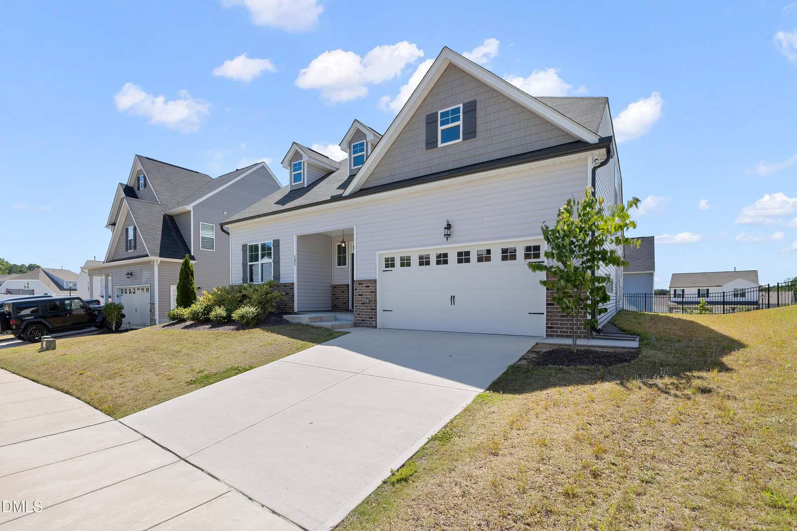 A modern two-story house with light-colored siding, a double garage, covered front porch, and a sloped driveway. The yard is mostly grass with a few small bushes and a tree. Blue sky with some clouds above.