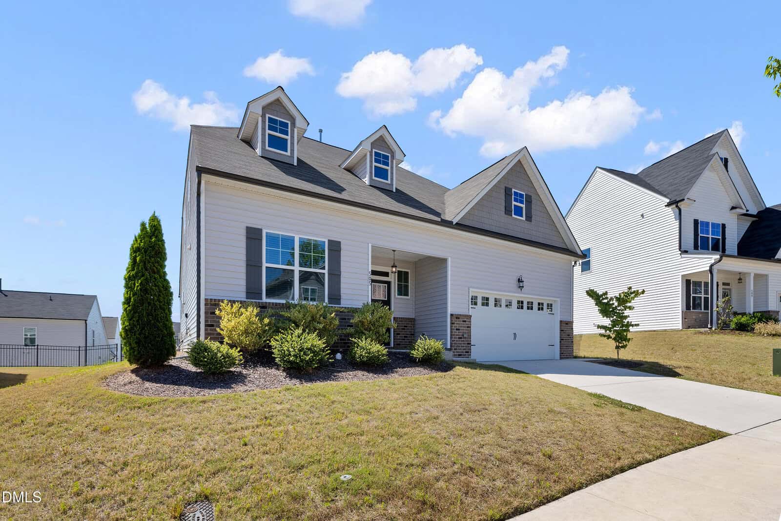 Two-story suburban house with gray roof, white siding, and two dormer windows. Attached double garage, manicured shrubs, and sloped front lawn under a blue sky with clouds.