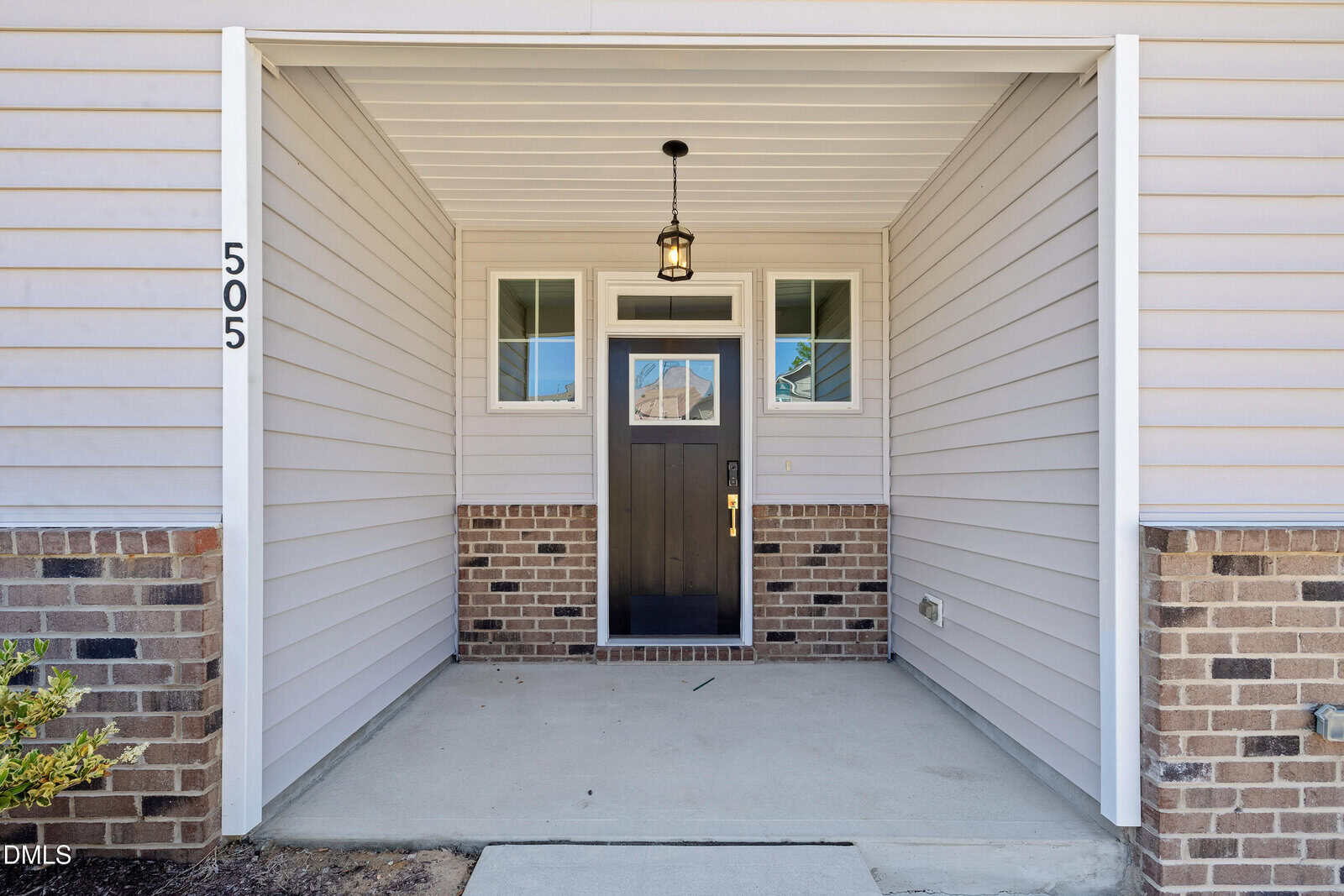 A covered front entryway with beige siding, brick accents, a brown door with glass panels, side windows, and the house number 505 mounted vertically on the left side. A hanging light fixture is above the doorway.