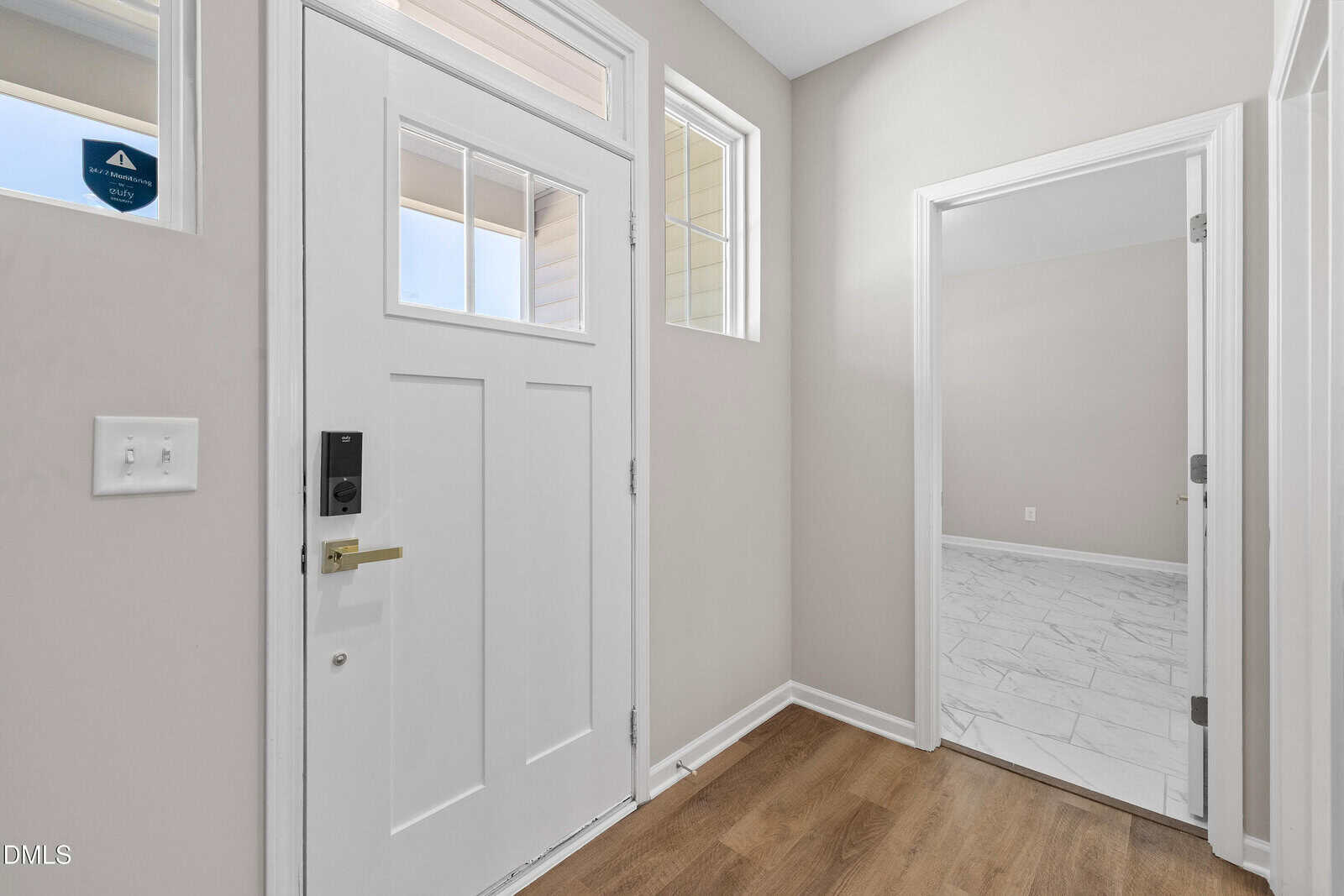 A white front door with windows is next to a light switch and beige walls. An open doorway leads to a room with white tile flooring. The floors in the entryway are light wood.