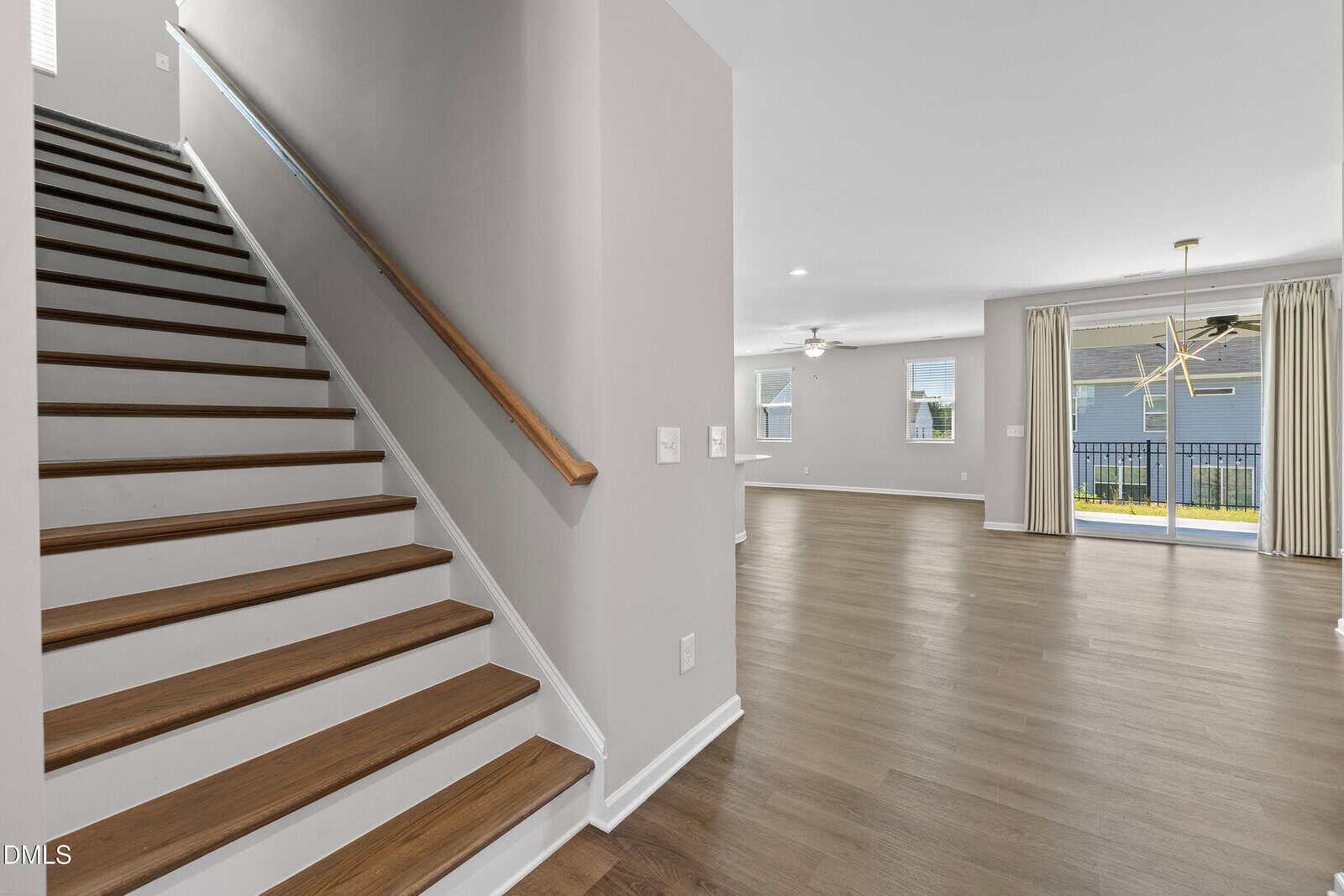 A modern, empty living space with wood floors, beige walls, a staircase with wooden steps, large windows, and sliding glass doors leading to a fenced yard.