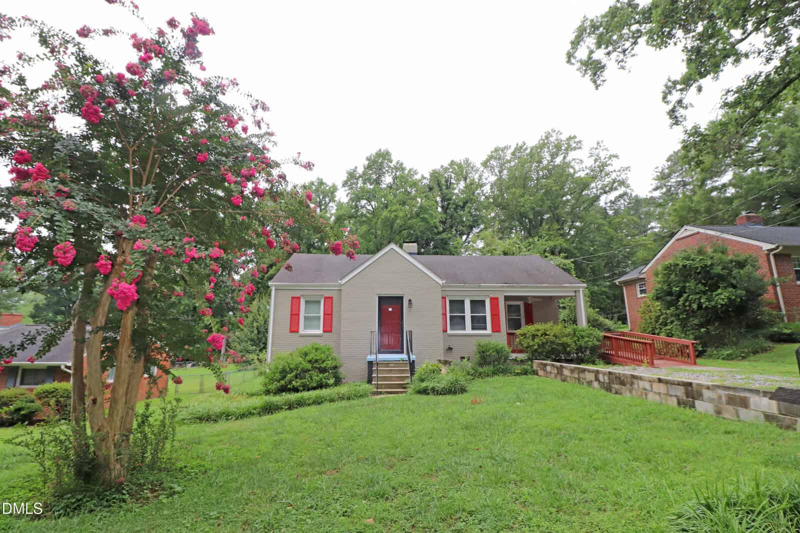 Single-story house at 132 Watson Drive, Henderson, with light-colored siding, red shutters, a front porch, and steps leading up. Pink flowering tree on the left, grass lawn, and driveway with a red railing on the right. Other houses and trees in the background.
