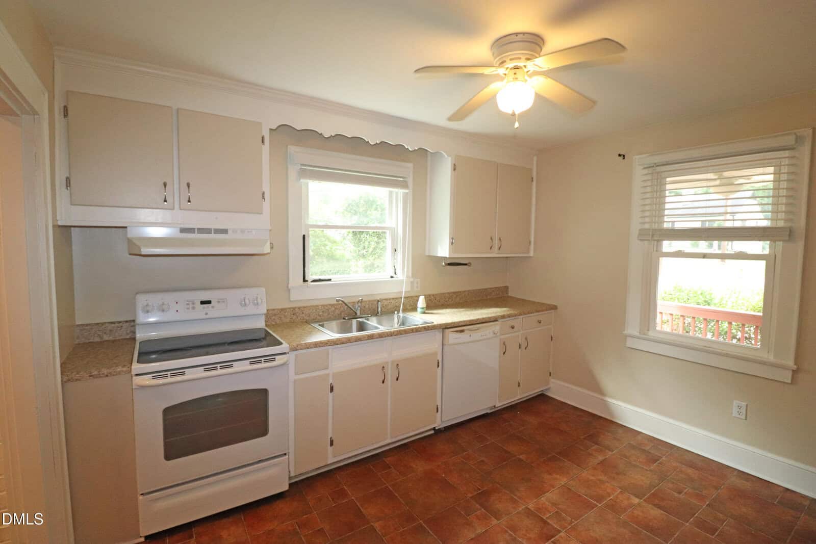 Bright kitchen with white cabinets, stove, dishwasher, double sink, beige countertops, tile floor, ceiling fan, and two windows letting in natural light.