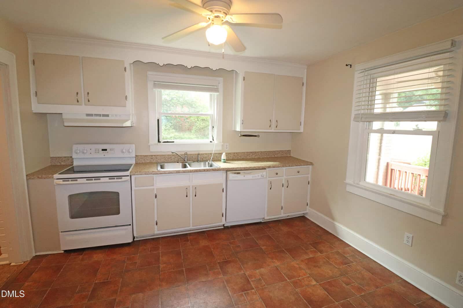 A kitchen with white cabinets, a white stove, dishwasher, double sink, tan countertops, tile flooring, a ceiling fan, and two windows with views of greenery and a porch.