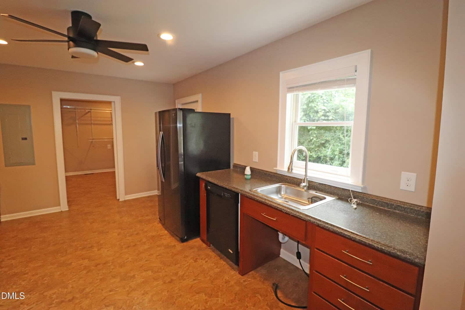 Kitchen with brown cabinets, black appliances, a sink under a window, and a ceiling fan. An open doorway leads to another room with built-in shelving. The walls are beige and floor is a wood-like material.