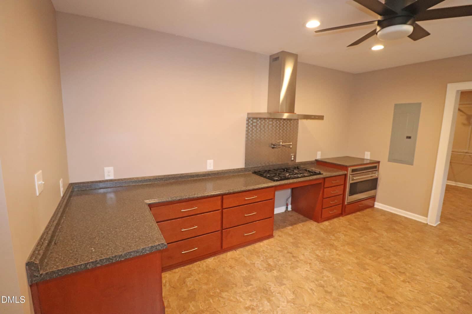 Modern kitchen with brown cabinets, black countertops, a stovetop with a stainless steel hood, built-in oven, and cork flooring. Neutral walls and ceiling lights complete the contemporary look.
