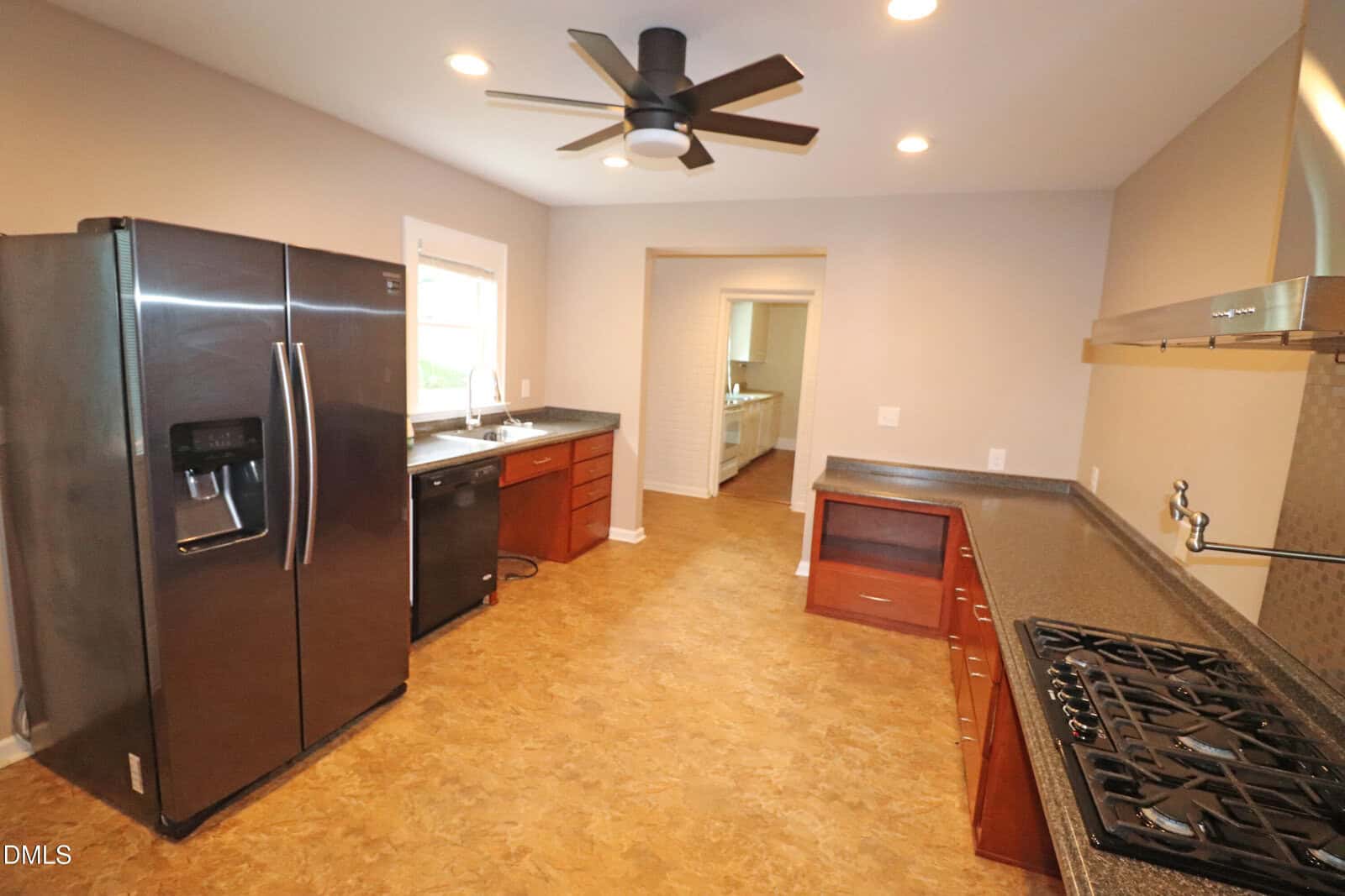 Modern kitchen with stainless steel appliances, dark countertops, wood cabinets, a ceiling fan, and recessed lighting. A window is above the sink, and an open doorway leads to another room.