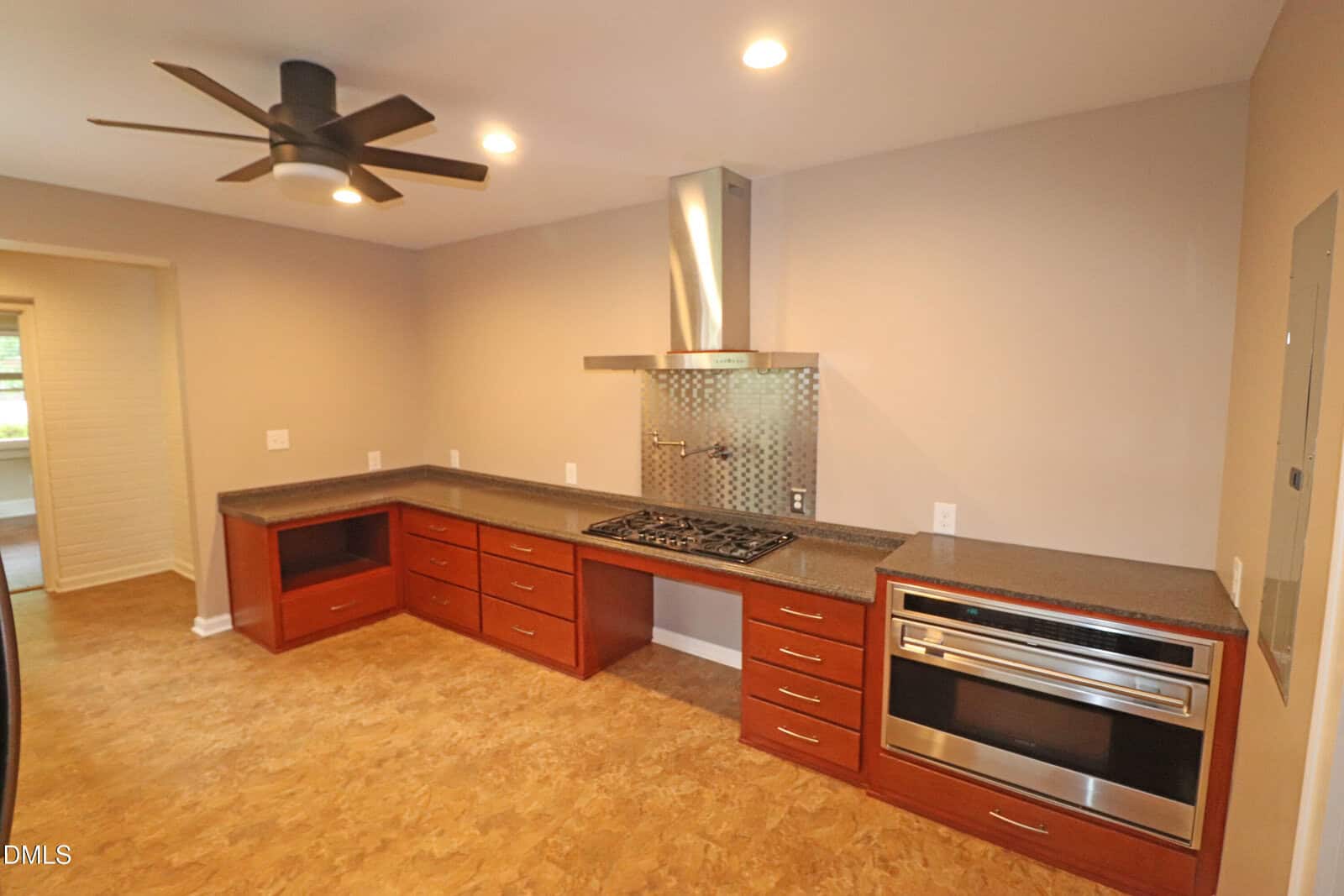 Modern kitchen with brown cabinets, black countertops, a built-in gas stove, stainless steel oven, vent hood, and ceiling fan. Room has beige walls and wood-patterned flooring. No visible decorations or appliances on counters.