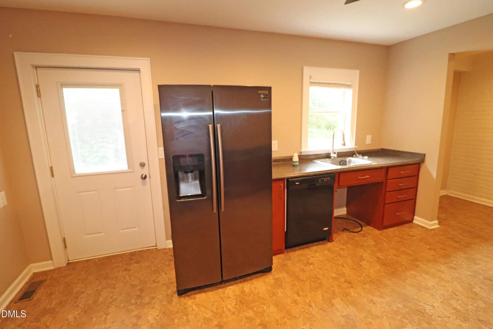 A kitchen with a side-by-side black refrigerator, a white door, a window above a sink, brown cabinets, a black dishwasher, granite countertop, and tan walls and floor.