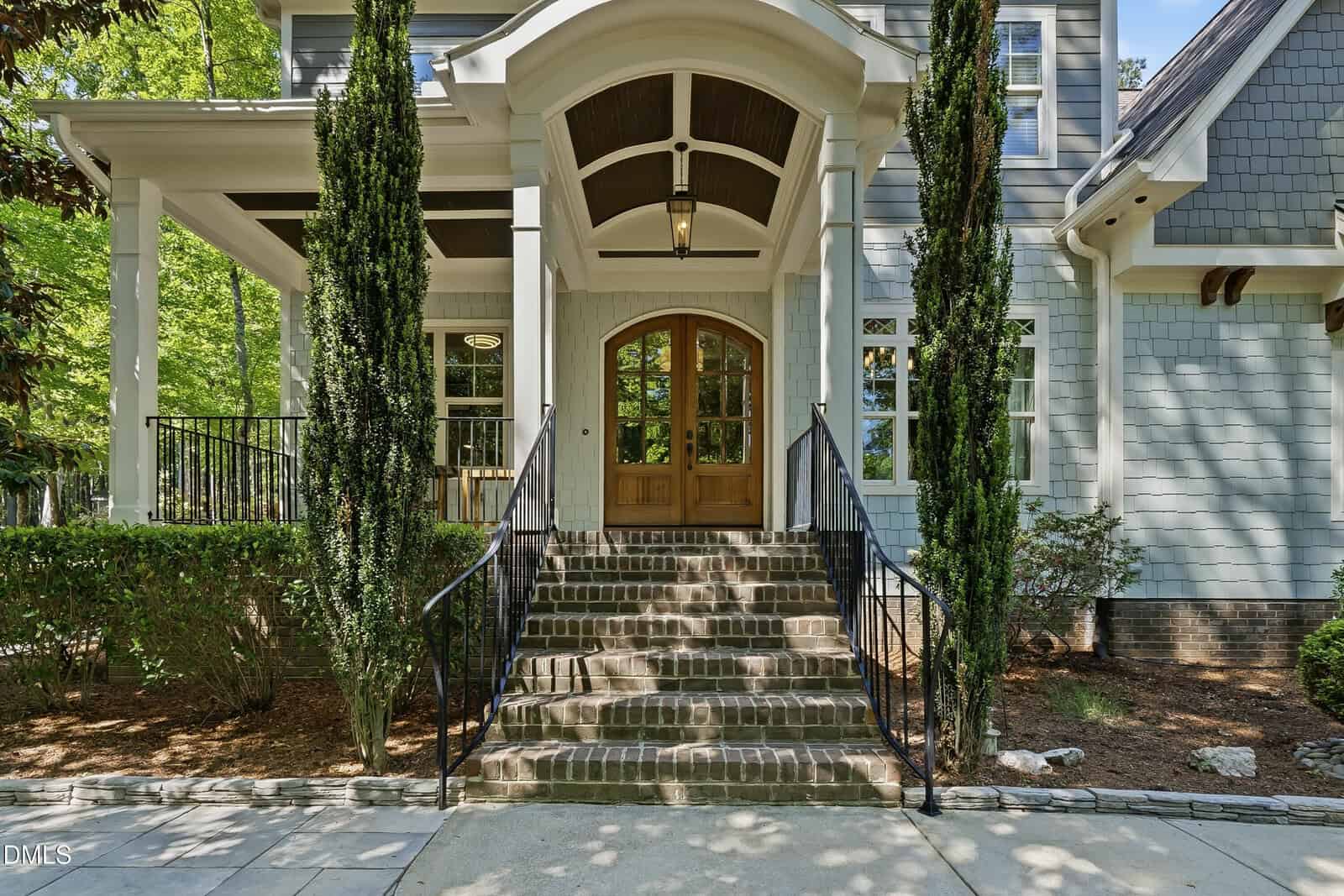 Front view of a house with gray siding, wooden double doors, large arched porch, brick steps, black railings, and tall evergreen shrubs flanking the entrance. Sunlight filters through surrounding trees.