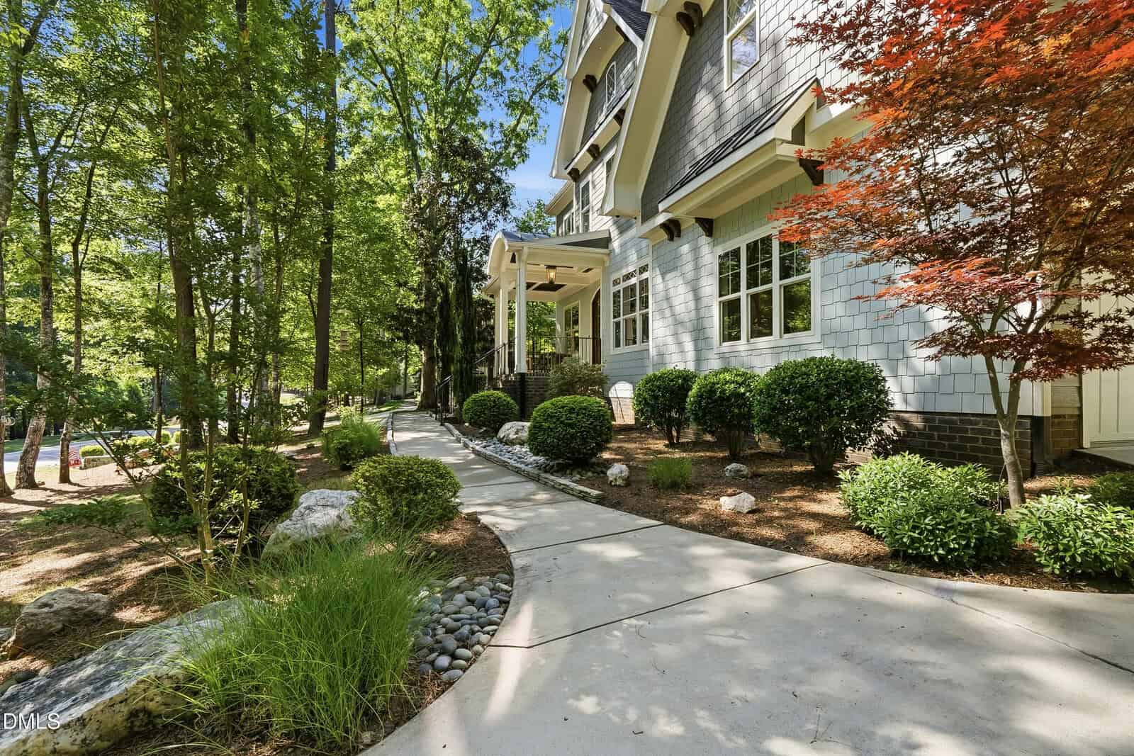 Curved concrete walkway leads to the front porch of a house with white siding, surrounded by bushes, small trees, and natural landscaping in a wooded area on a sunny day.