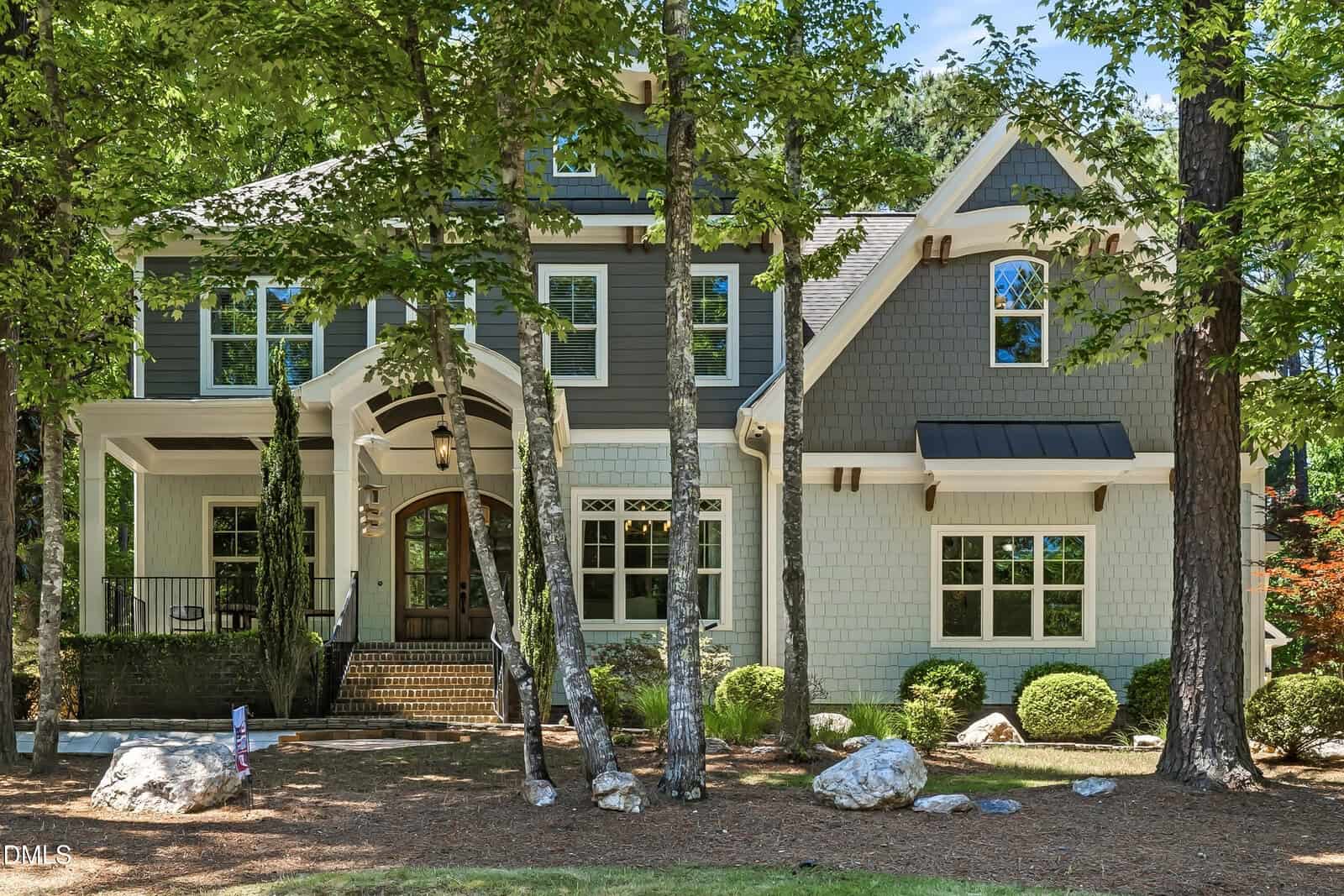 Two-story gray and white house with a covered front porch, arched entryway, large windows, and surrounded by trees and landscaping at 3576 Bragg Valley Lane in Wake Forest. Rocks are scattered on the front lawn.