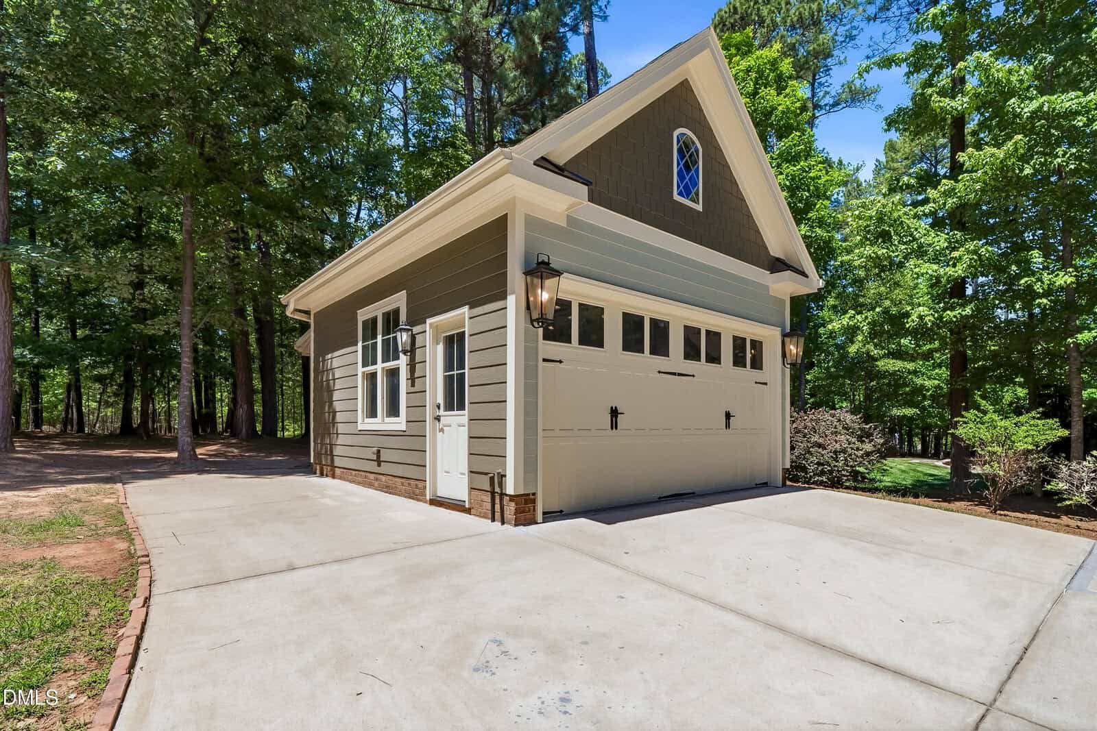 Detached single-car garage with taupe siding, white trim, and carriage-style door, surrounded by trees and a concrete driveway on a sunny day.