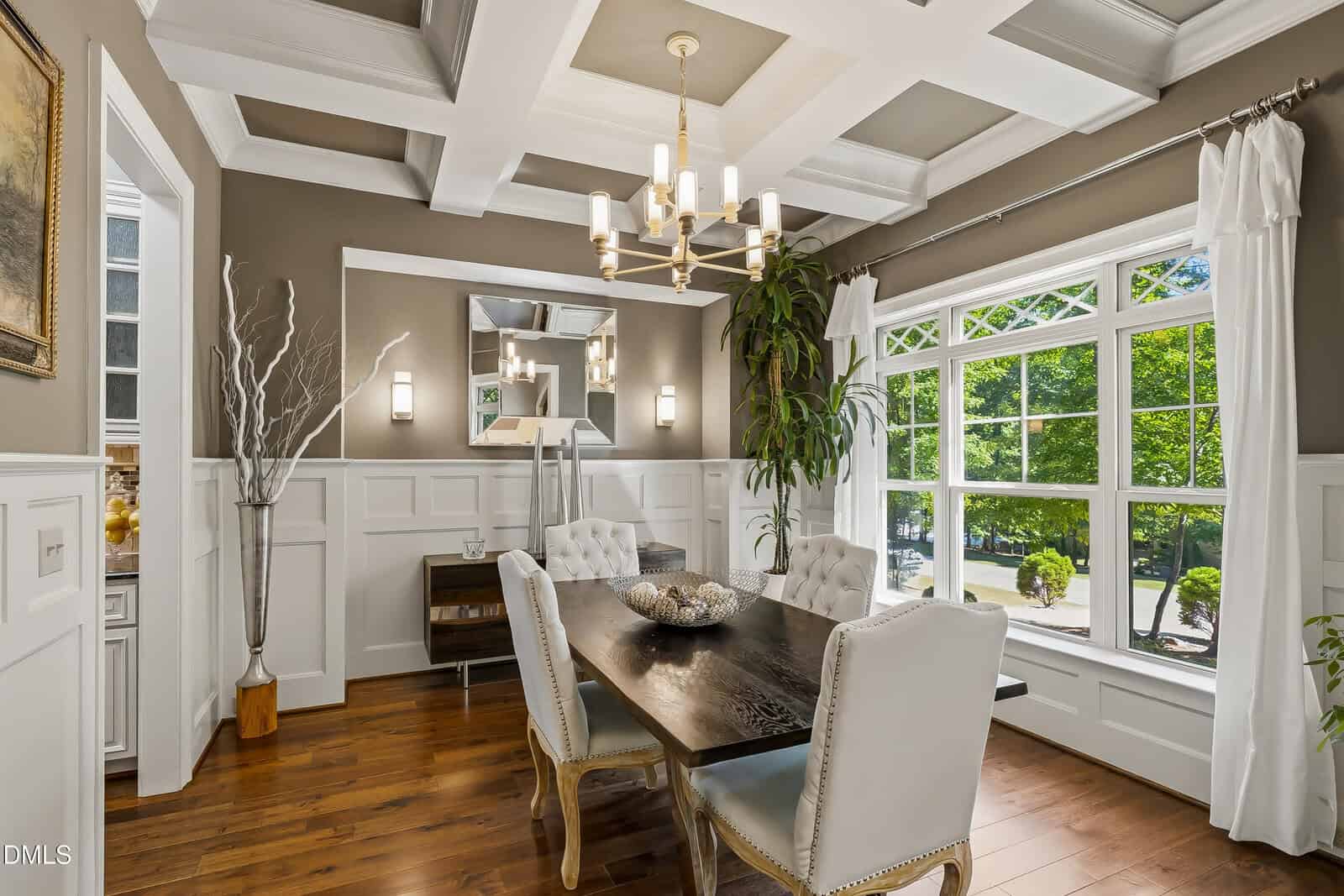 Elegant dining room with a dark wood table, tufted white chairs, chandelier, large windows with a view of greenery, white wainscoting, wood floors, and decorative plants.