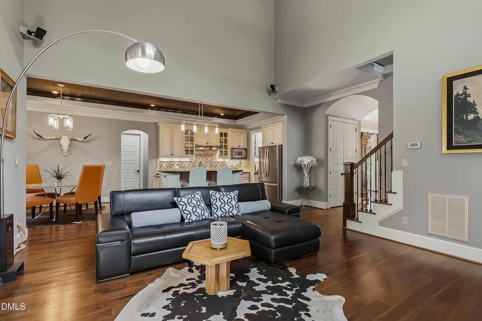 Modern living room with black sectional, wooden coffee table, cowhide rug, and open view into a kitchen with white cabinets and pendant lights. A dining area with orange chairs and decorative deer head is visible on the left.