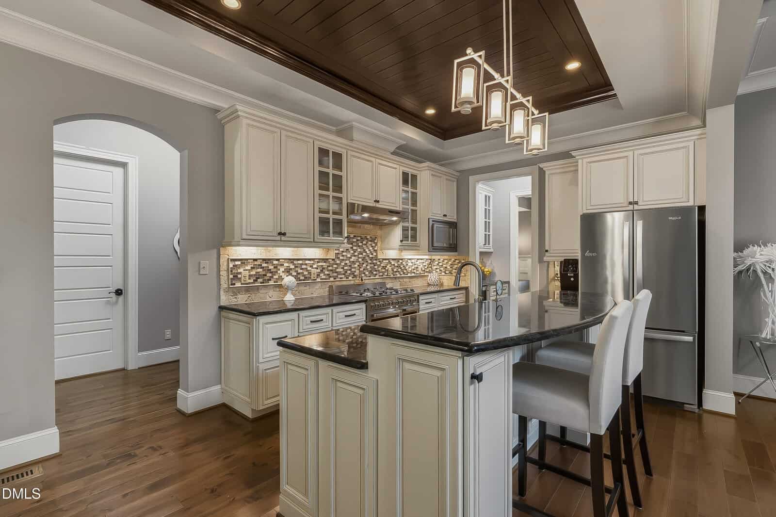 Modern kitchen with white cabinets, stainless steel appliances, mosaic tile backsplash, dark wood ceiling accents, pendant lighting, a black granite island, and two white barstools on polished wooden floors.