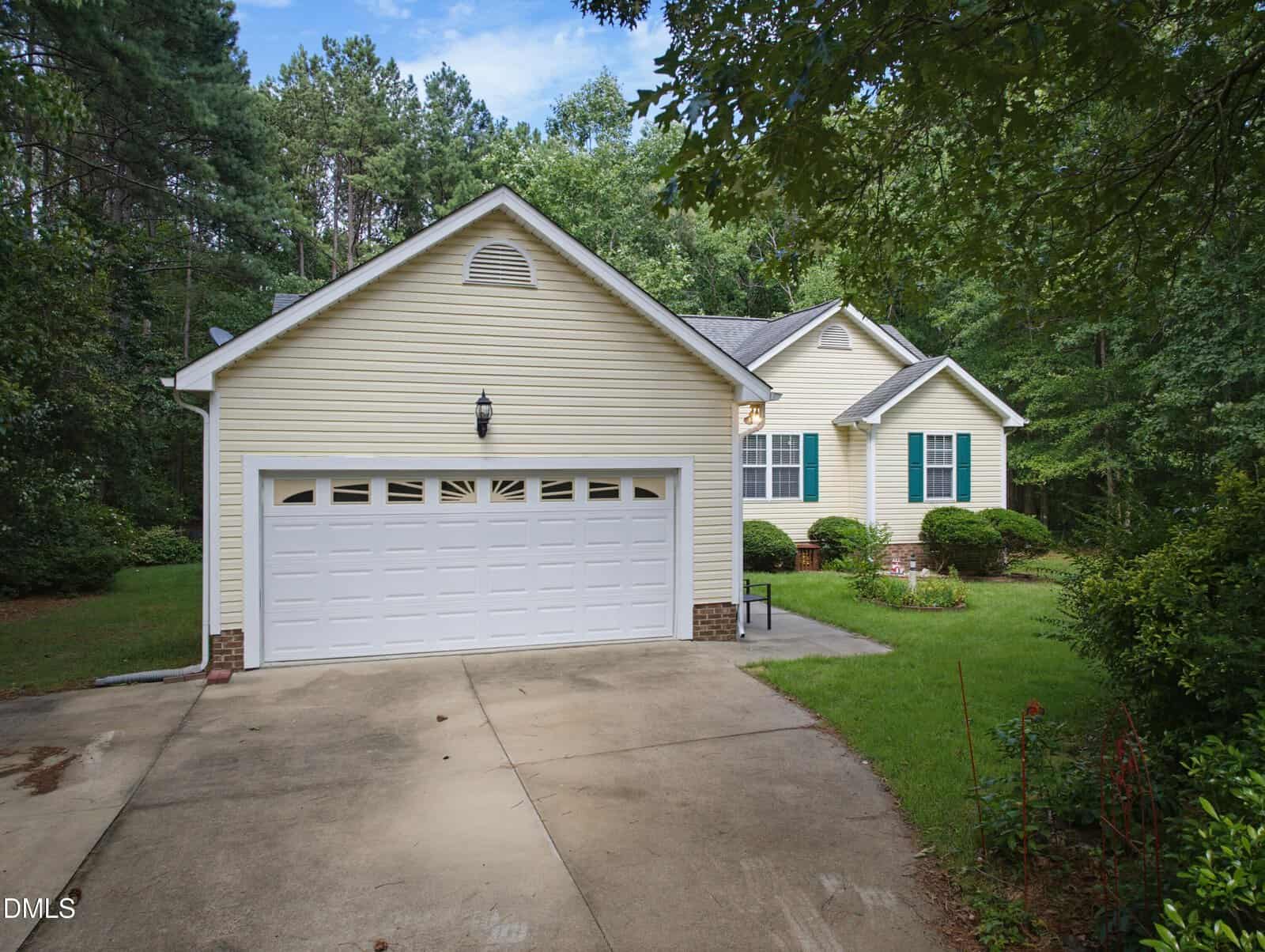 Single-story yellow house with white trim and a double garage, set among trees with a concrete driveway and a small, manicured front yard.